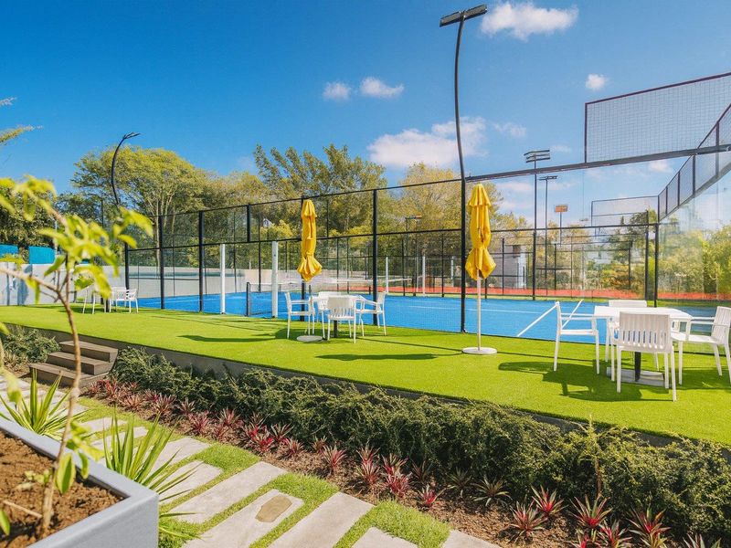 Well-maintained outdoor sports facility featuring a blue padel court with perimeter fencing, artificial turf viewing area, and integrated landscaping.