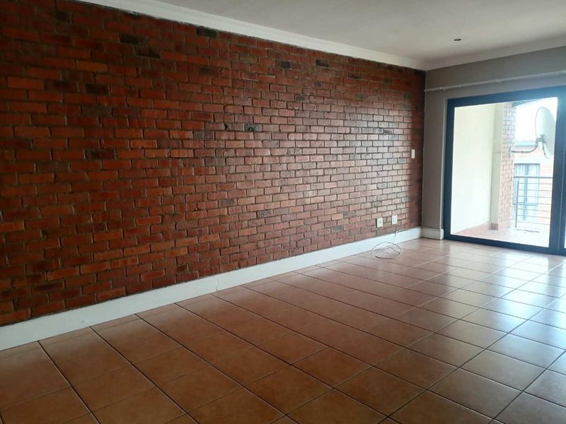 An empty living room featuring a prominent exposed brick wall, ceramic tiled flooring, and a sliding glass door leading to an outdoor balcony.