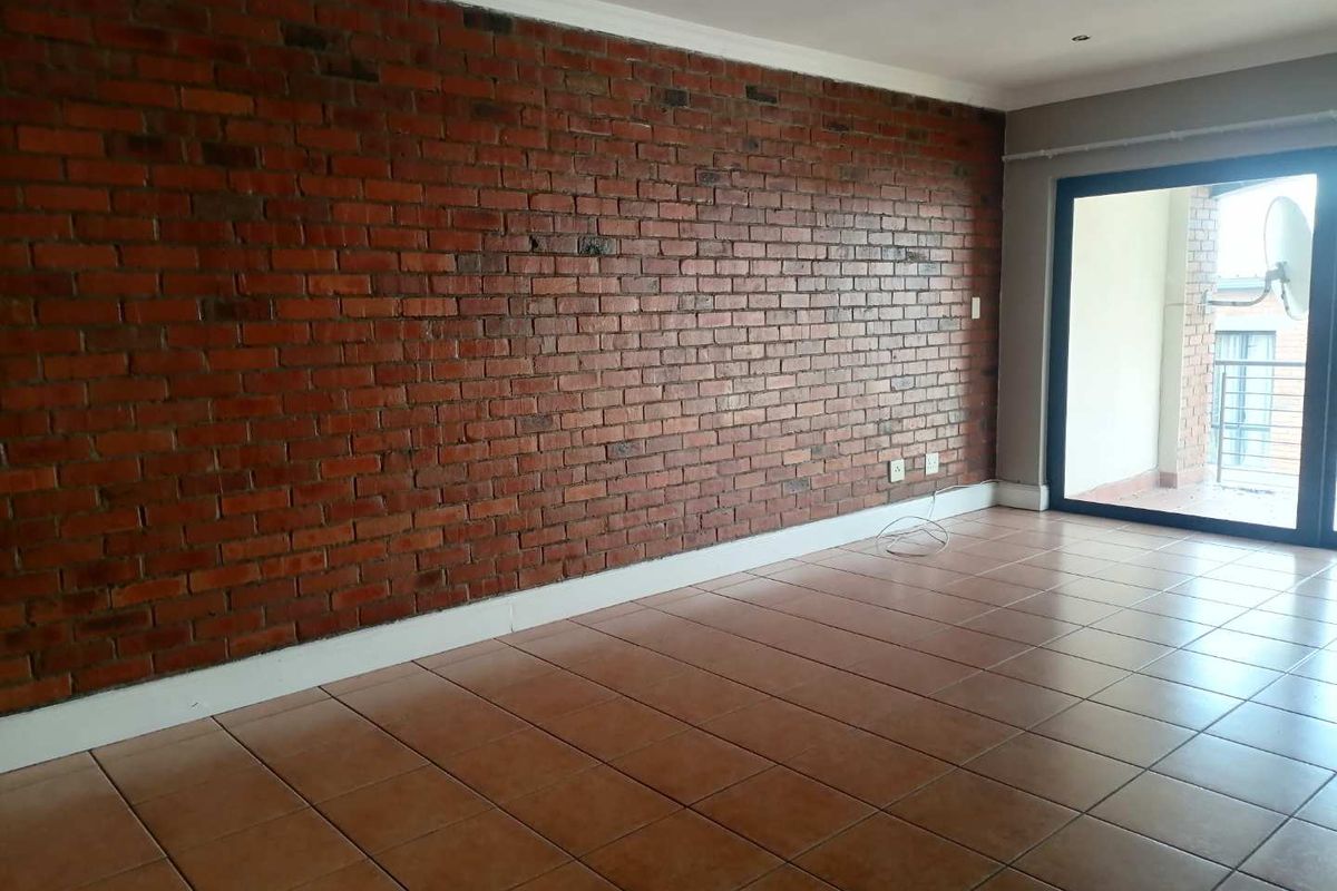 An empty living room featuring a prominent exposed brick wall, ceramic tiled flooring, and a sliding glass door leading to an outdoor balcony.