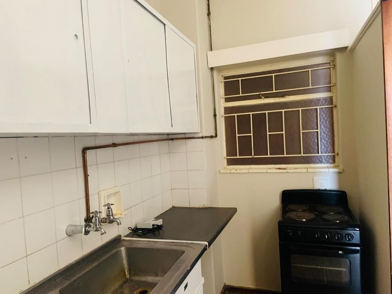 Compact kitchen featuring a stainless steel sink, tiled splashback, upper cabinetry, and a freestanding stove with exposed plumbing and security bars on the window.