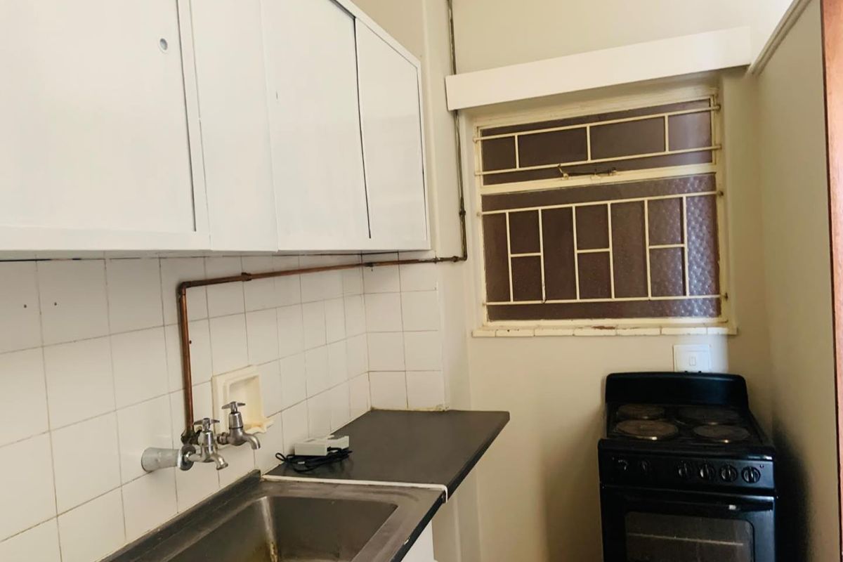 Compact kitchen featuring a stainless steel sink, tiled splashback, upper cabinetry, and a freestanding stove with exposed plumbing and security bars on the window.