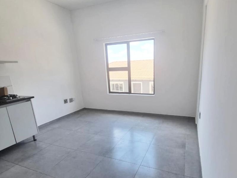 An empty open-plan living area featuring modern grey floor tiles, a compact kitchen unit with a built-in hob, and neutral white walls.