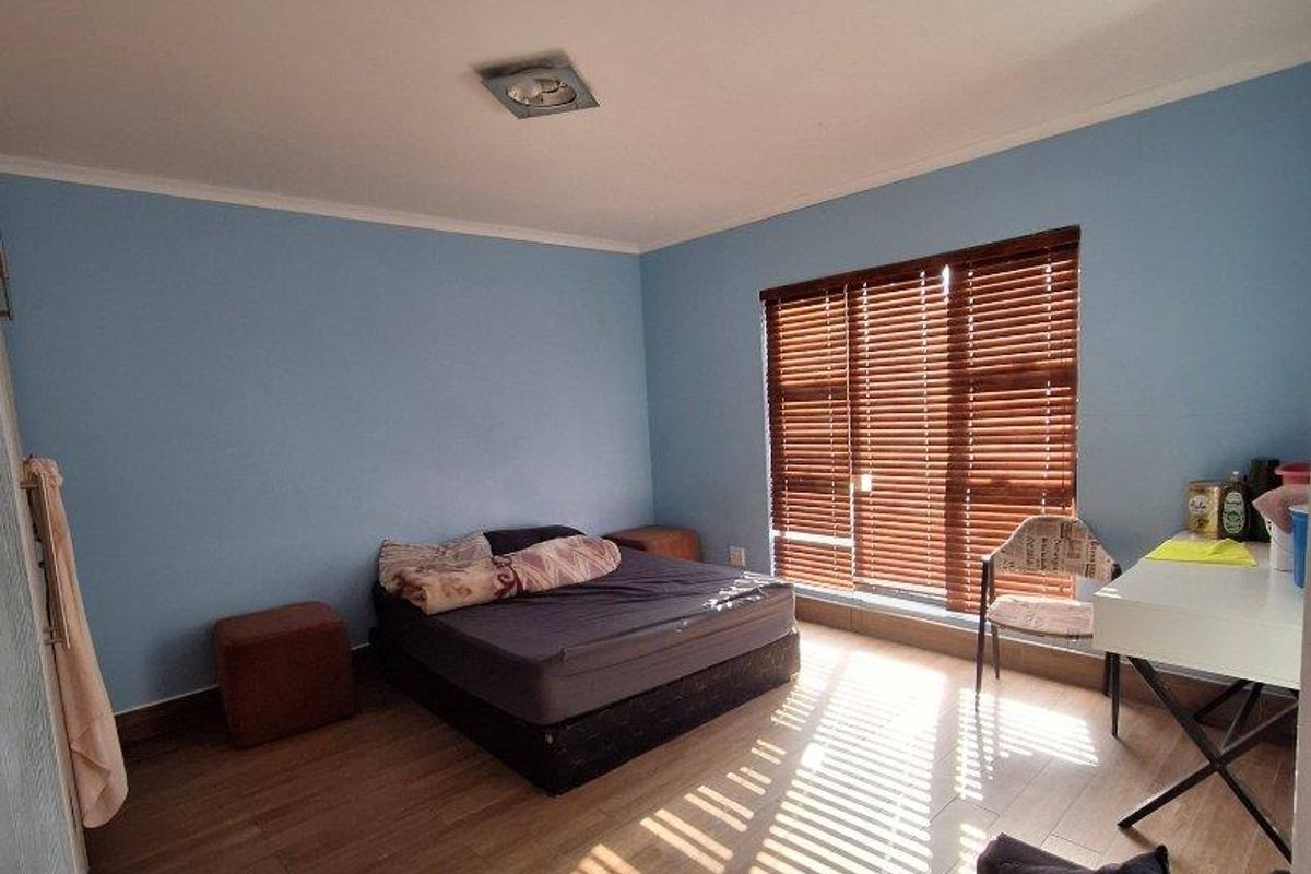 A spacious bedroom featuring wood-look tiled flooring, light blue walls, and large windows fitted with wooden venetian blinds.