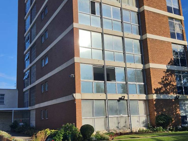 A view of the apartment building facade featuring brick construction, large windows, and well-maintained surrounding garden beds.