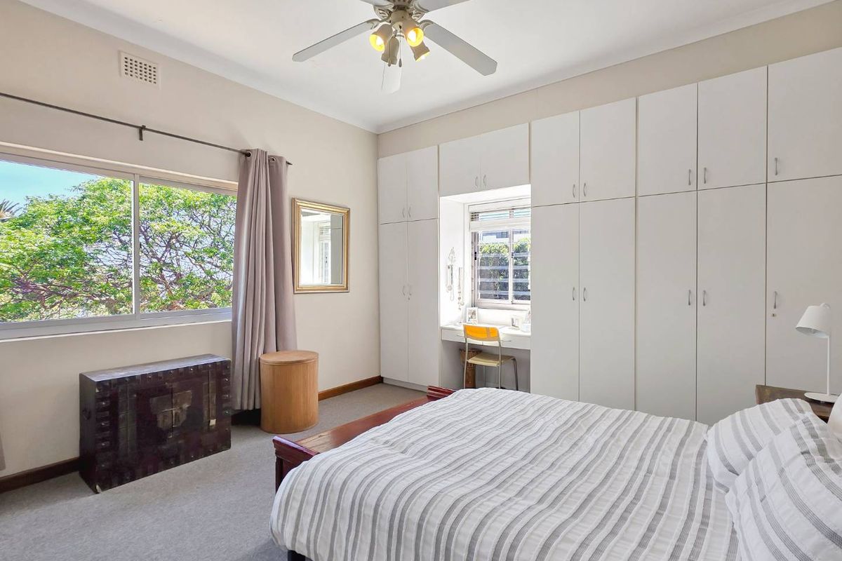 A well-lit bedroom featuring extensive built-in white cabinetry, a integrated desk area, ceiling fan, and large windows overlooking greenery.