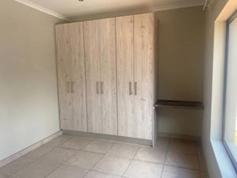 A clean, empty bedroom featuring light wood-finish built-in cupboards, an integrated desk area, and neutral tiled flooring.