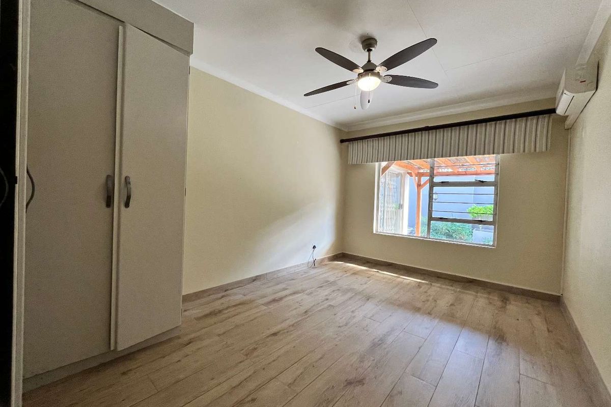 A bright, empty bedroom featuring laminate flooring, built-in cupboards, a ceiling fan, and an air conditioning unit.