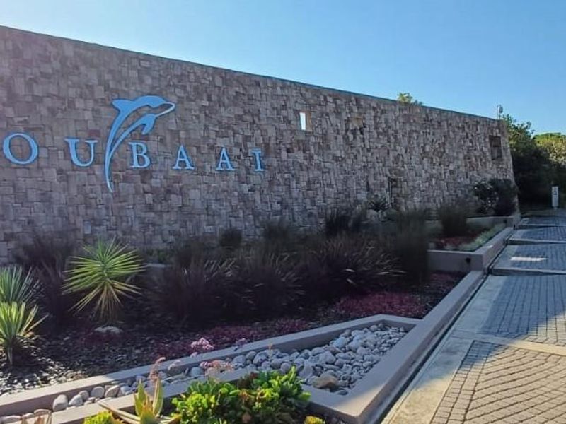 The entrance to Oubaai estate featuring a prominent stone-clad boundary wall, paved access road, and well-maintained landscaped garden beds.