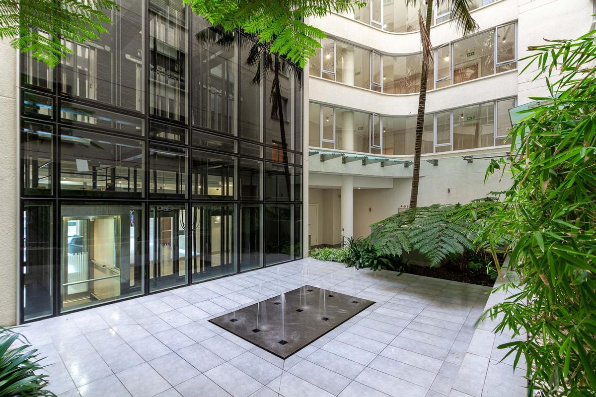 A modern apartment complex courtyard featuring a central water feature, tiled flooring, glass facade, and landscaped greenery.