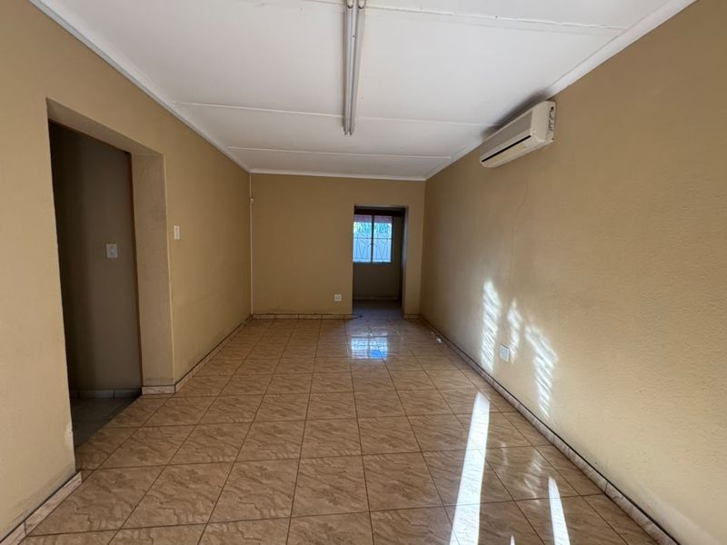 An empty, neutrally painted living area featuring tiled flooring, a wall-mounted air conditioning unit, and direct access to adjacent rooms.