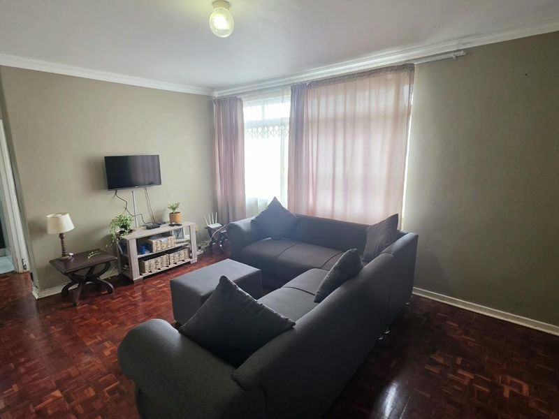 A living room featuring traditional parquet flooring, coved ceilings, and large windows with curtain tracks, offering a functional space with potential for cosmetic updates.