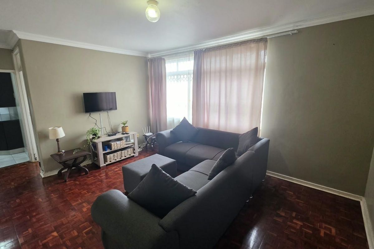 A living room featuring traditional parquet flooring, coved ceilings, and large windows with curtain tracks, offering a functional space with potential for cosmetic updates.