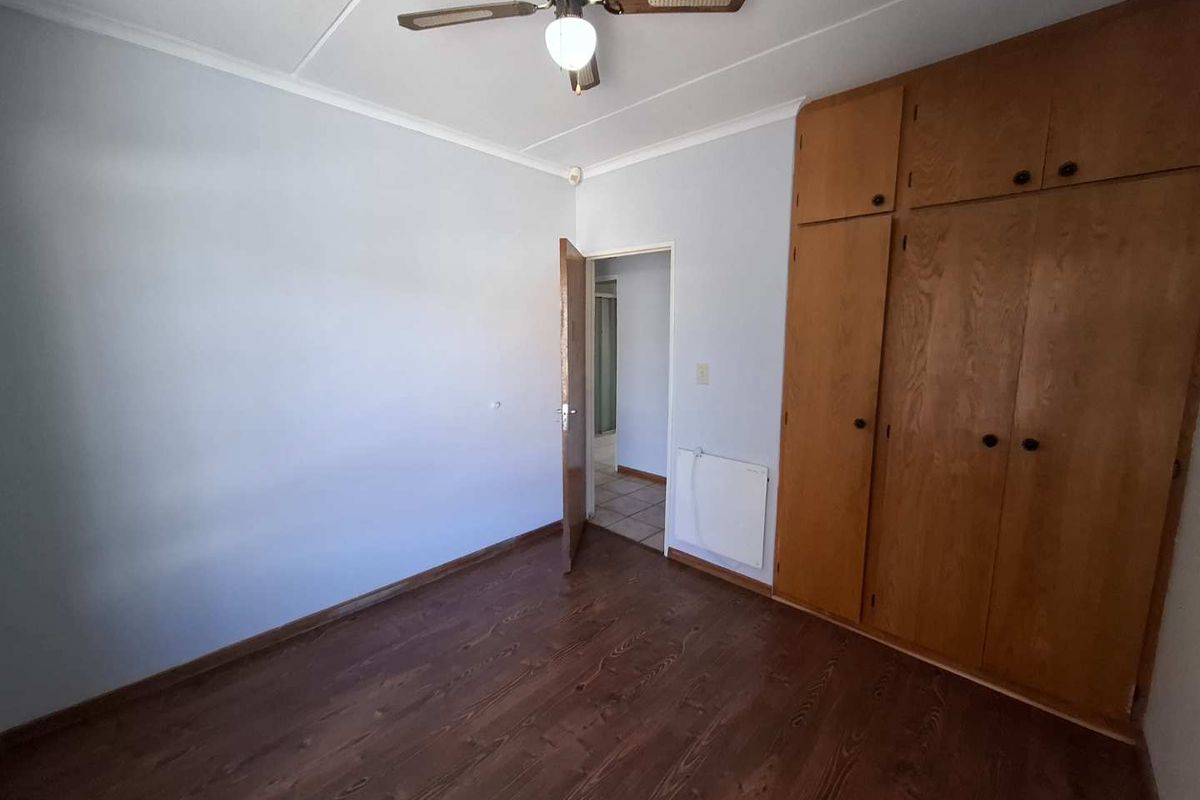 A bedroom featuring built-in wooden cupboards, a ceiling fan, and wood-look flooring, with an adjacent doorway leading to another room.