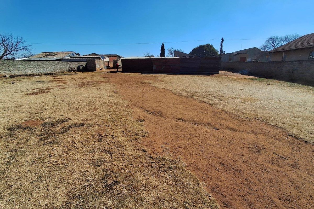 A view of a vacant residential plot featuring a large open yard area, perimeter brick walling, and an existing outbuilding structure.