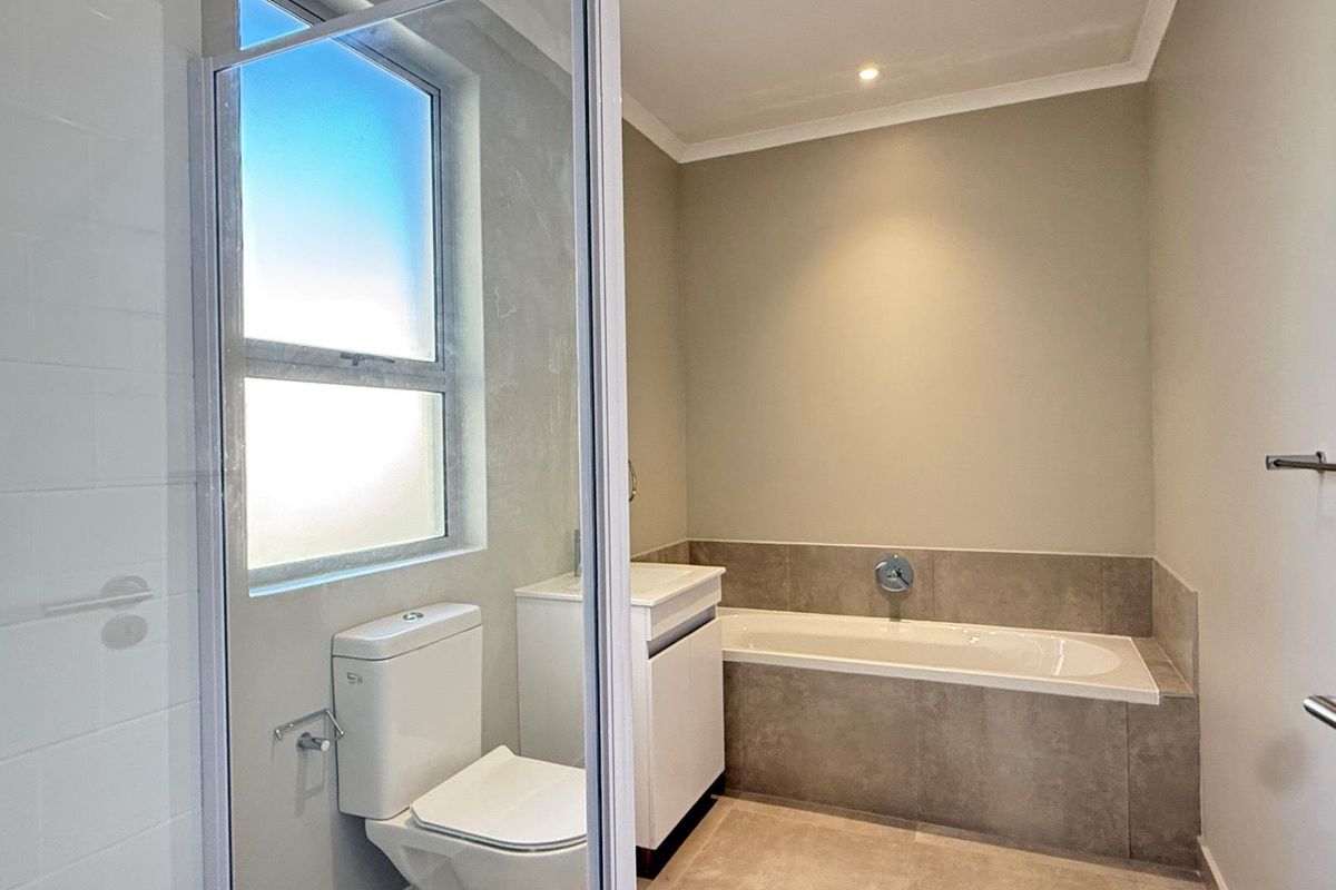 A modern, clean bathroom featuring a built-in bathtub, a vanity cabinet, a toilet, and a glass-enclosed shower with neutral tiled finishes.