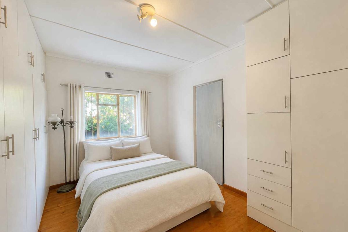 A bright bedroom featuring polished timber flooring, extensive built-in cabinetry, and a window with security bars overlooking the garden.