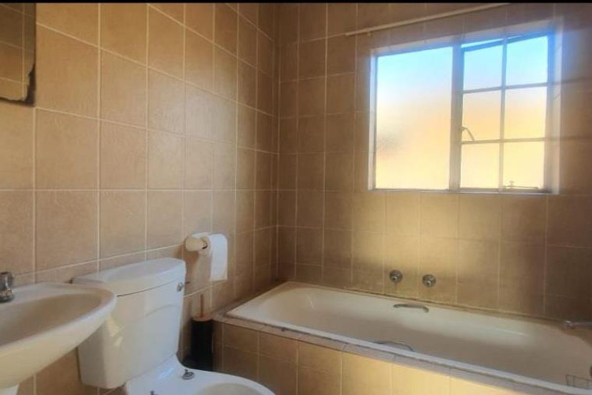 A functional bathroom featuring a built-in bathtub, pedestal basin, and toilet, finished with neutral wall tiling and a window for natural light.