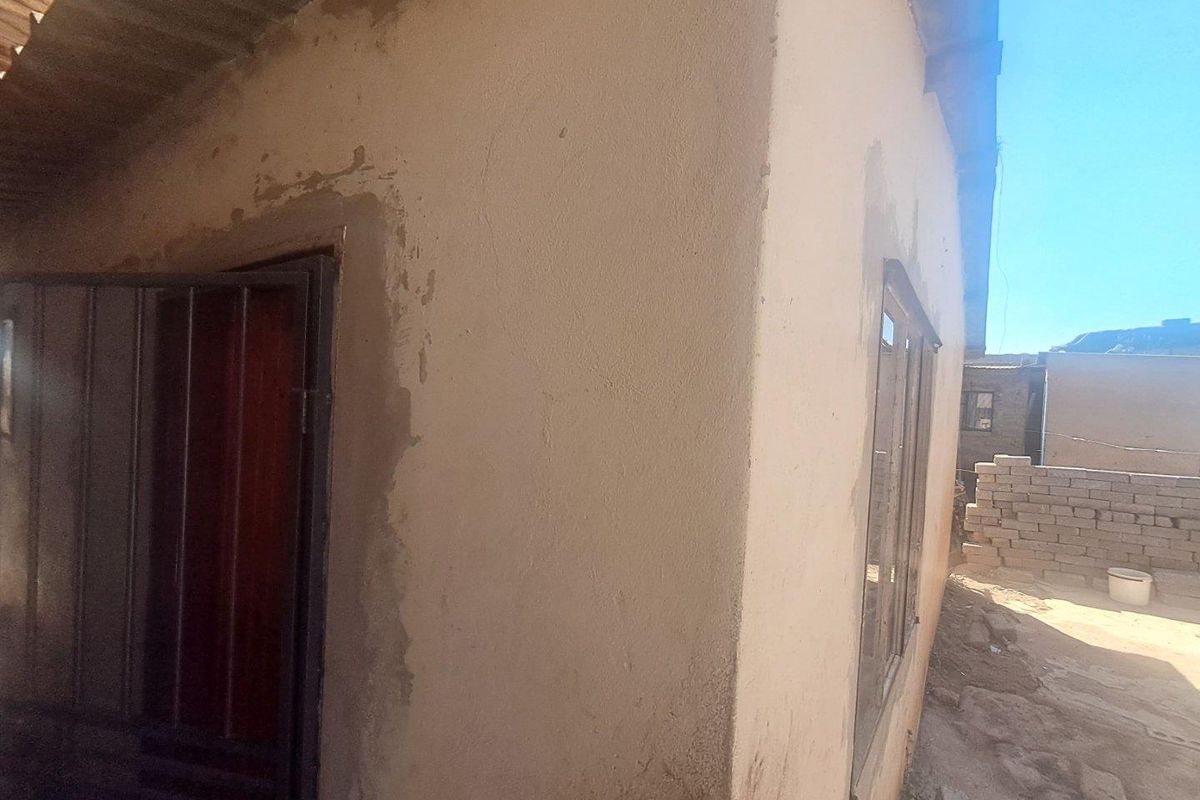 Exterior view of the property showing plastered masonry walls, security gate, and metal window frames with unfinished plastering work around the openings.