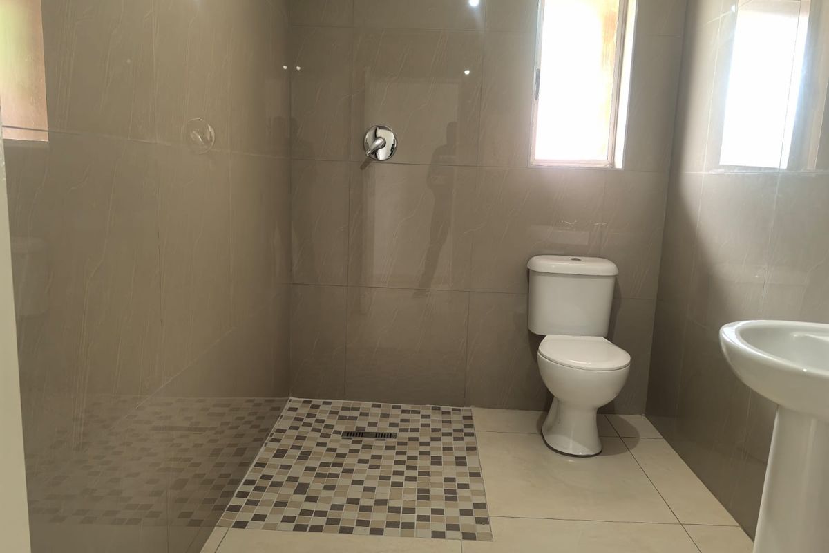 A clean, modern bathroom featuring a walk-in shower with mosaic floor detailing, a pedestal basin, and a standard toilet with neutral wall tiling.