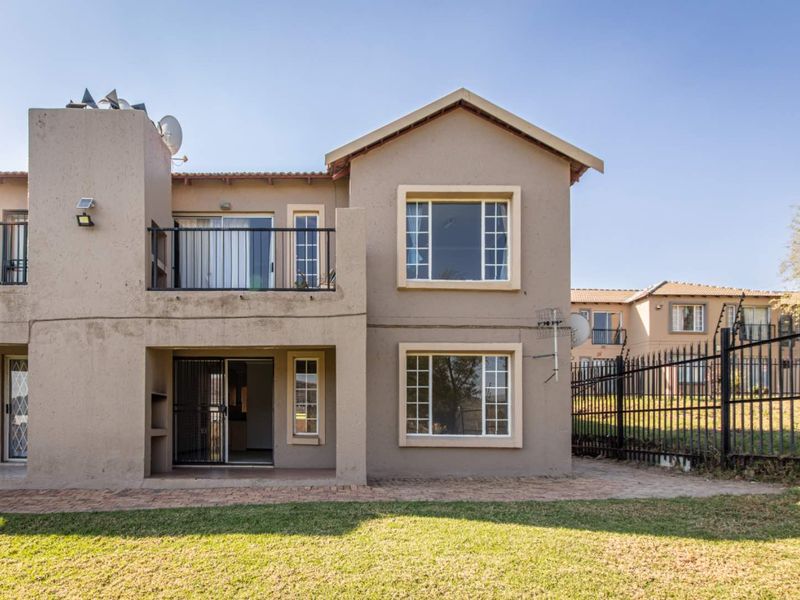 Rear view of a two-story apartment unit featuring a ground-floor patio, upper-level balcony, and paved garden area with perimeter security fencing.
