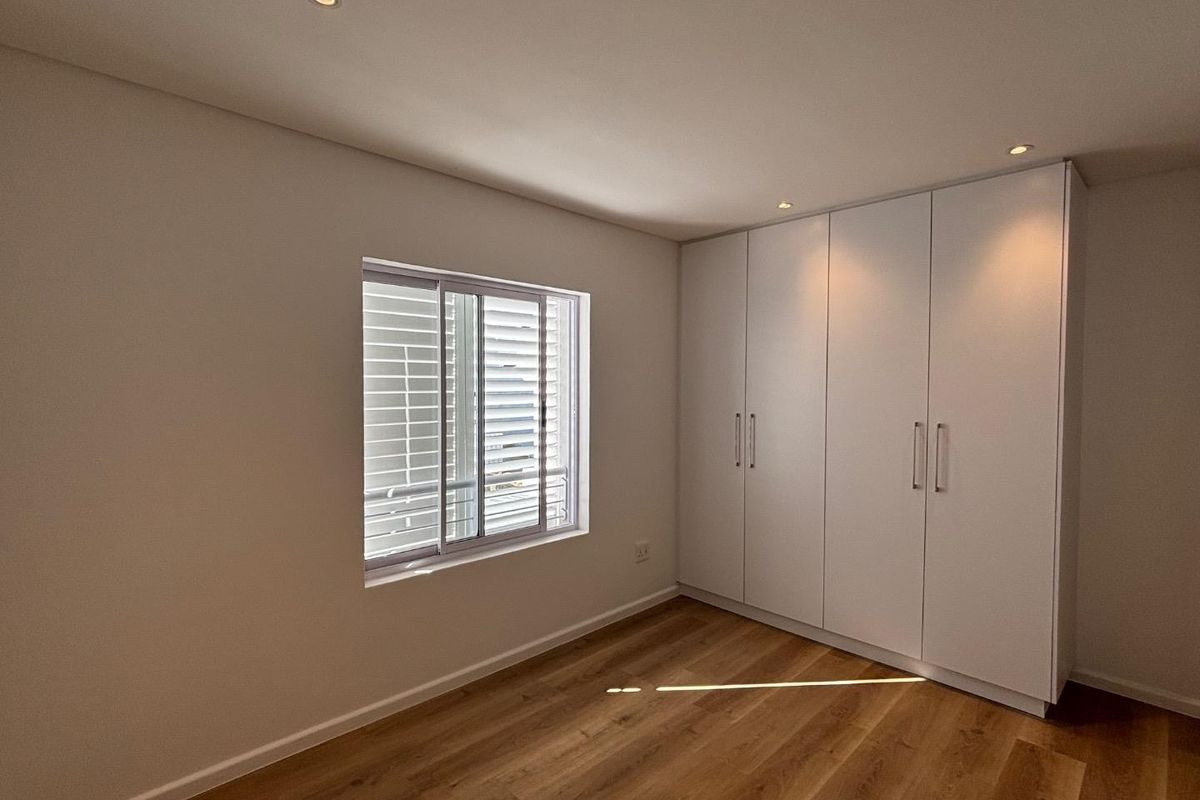 A bright, modern bedroom featuring light wood-look flooring, recessed ceiling lighting, and built-in floor-to-ceiling white cabinetry.