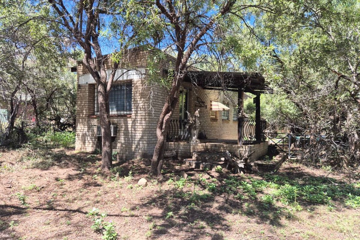 A rustic brick cottage exterior nestled in a wooded area, featuring a covered patio and security bars on the windows.