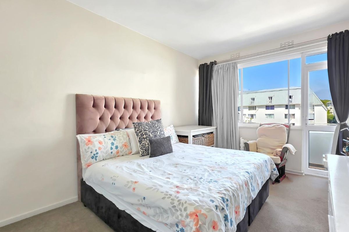 A well-lit bedroom featuring neutral walls, carpeted flooring, and a large window with access to a private balcony.