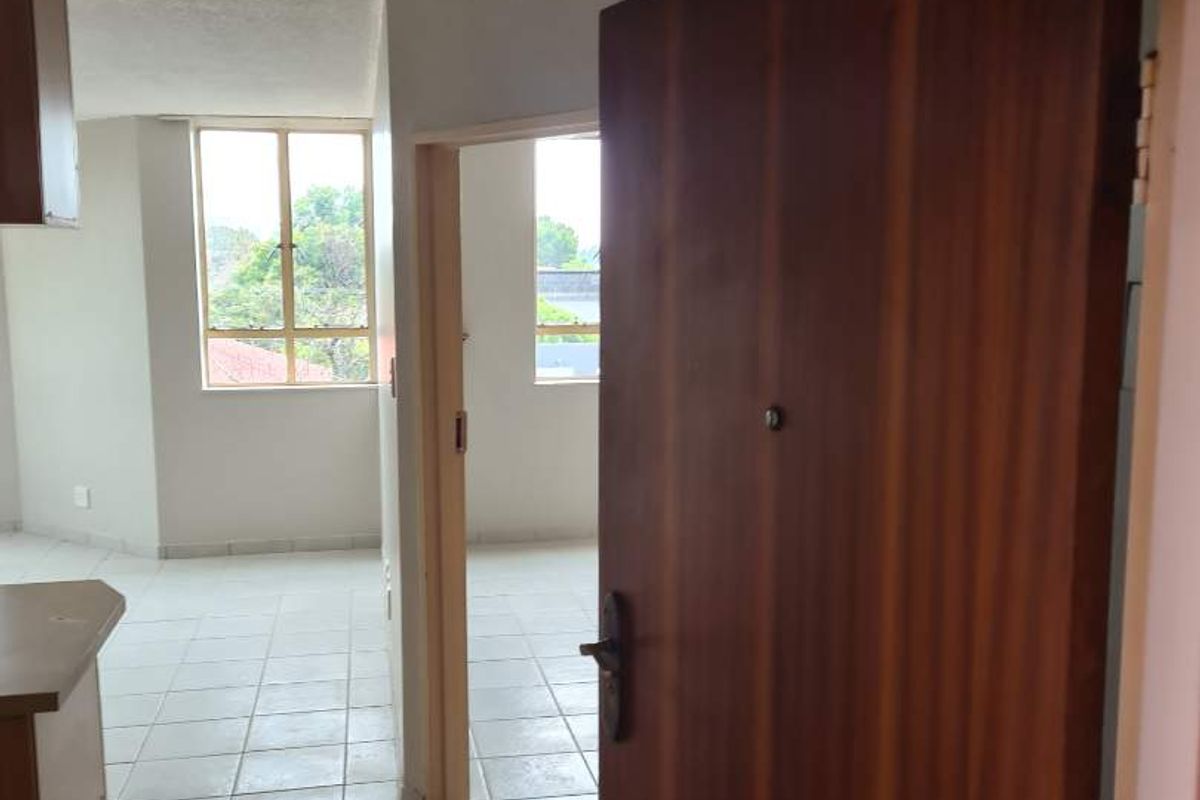An open-plan apartment interior featuring tiled flooring and a wooden entrance door, leading into a bright living and kitchen area.