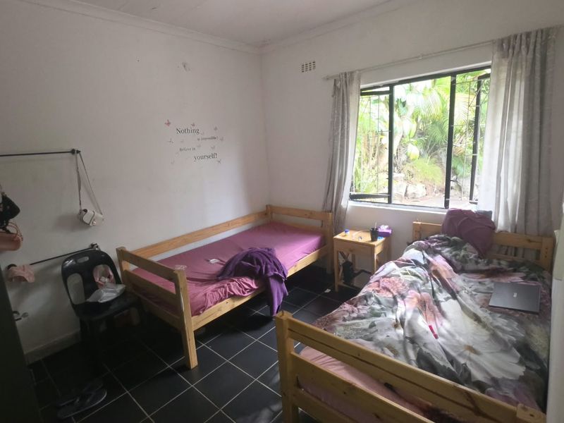 A bedroom featuring dark tiled flooring and a large window overlooking the garden, with visible wall scuffs and exposed electrical wiring.