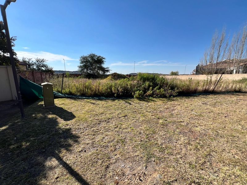 A vacant residential plot featuring a grassed area, perimeter walling, and an electrical utility box, with overgrown vegetation present.