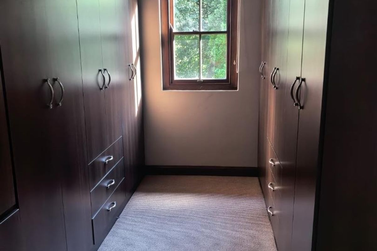 A walk-in dressing room featuring extensive dark wood built-in cabinetry, neutral carpeting, and a central window providing natural light.