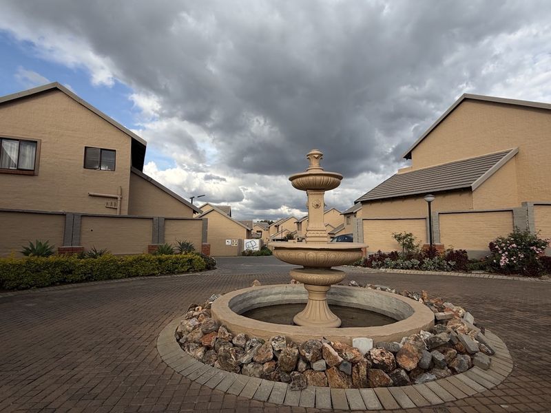 A well-maintained townhouse complex entrance featuring a central decorative water fountain, paved driveway, and established boundary walls under a cloudy sky.