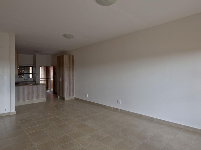 An open-plan living and kitchen area featuring neutral tiled flooring, built-in storage cupboards, and functional kitchen cabinetry in a clean, vacant space.