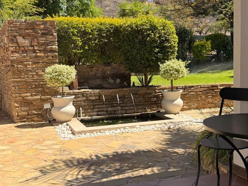 A covered patio area featuring stone paving, overlooking a landscaped garden with a built-in stone water feature and retaining wall.