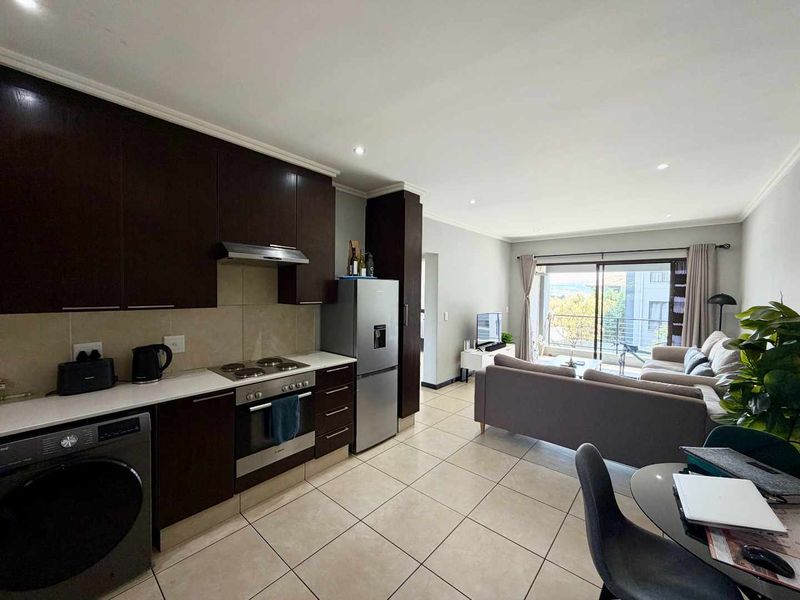 Open-plan living area featuring a modern kitchen with dark cabinetry, built-in appliances, and tiled flooring, flowing seamlessly into a bright lounge with balcony access.