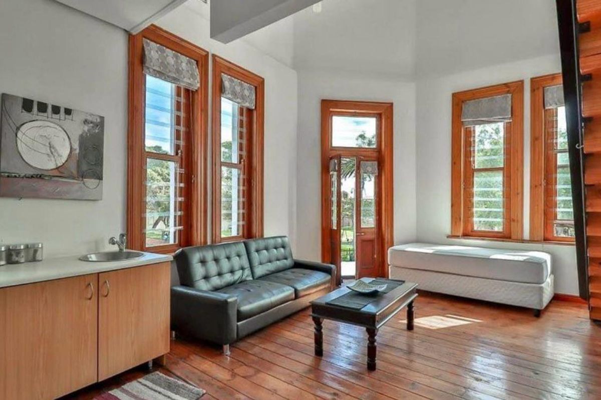 A bright living area featuring warm timber flooring, natural wood window frames, and a convenient built-in kitchenette with a sink.