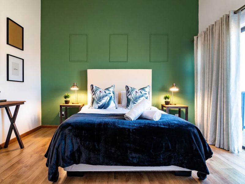 A well-lit bedroom featuring laminate flooring, a green accent wall with decorative molding, and a sliding glass door providing access to an outdoor area.