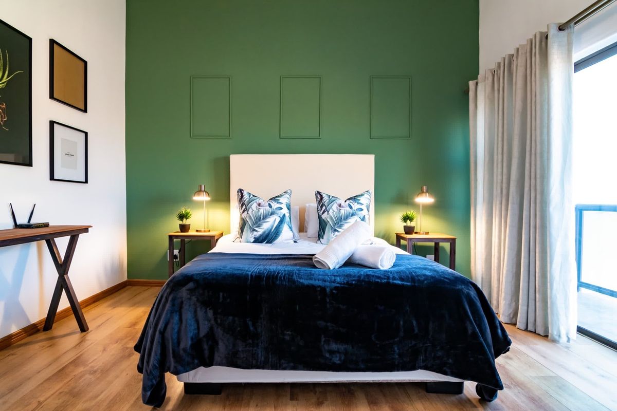 A well-lit bedroom featuring laminate flooring, a green accent wall with decorative molding, and a sliding glass door providing access to an outdoor area.