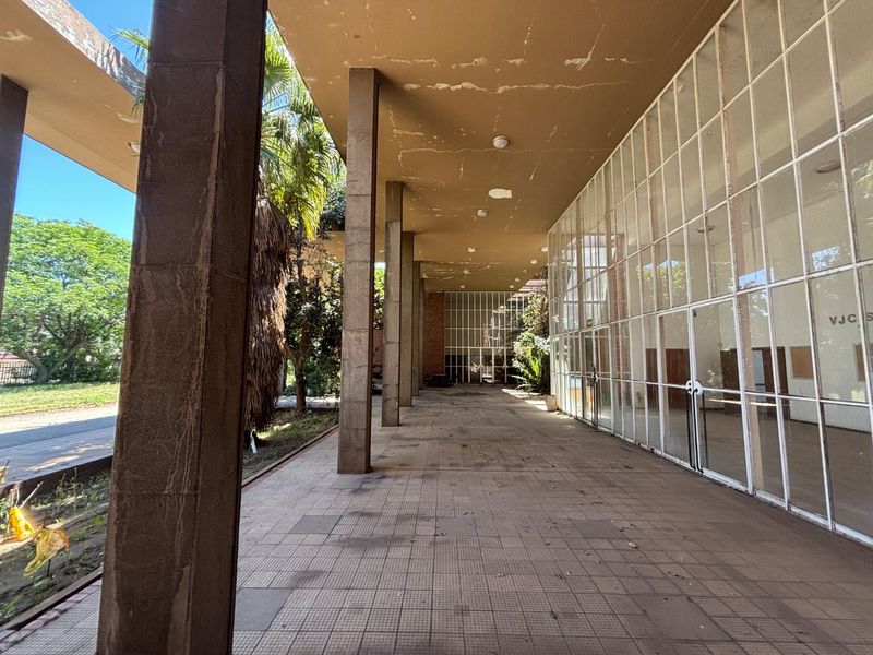 A spacious covered outdoor walkway featuring concrete columns, tiled flooring, and an extensive glass facade, showing signs of wear and requiring maintenance.
