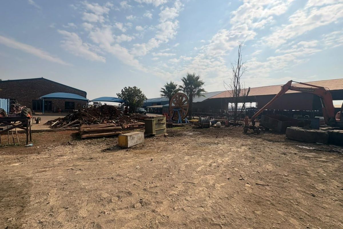 An industrial yard area featuring large metal-roofed sheds, brick structures, and open unpaved ground suitable for storage or operational use.