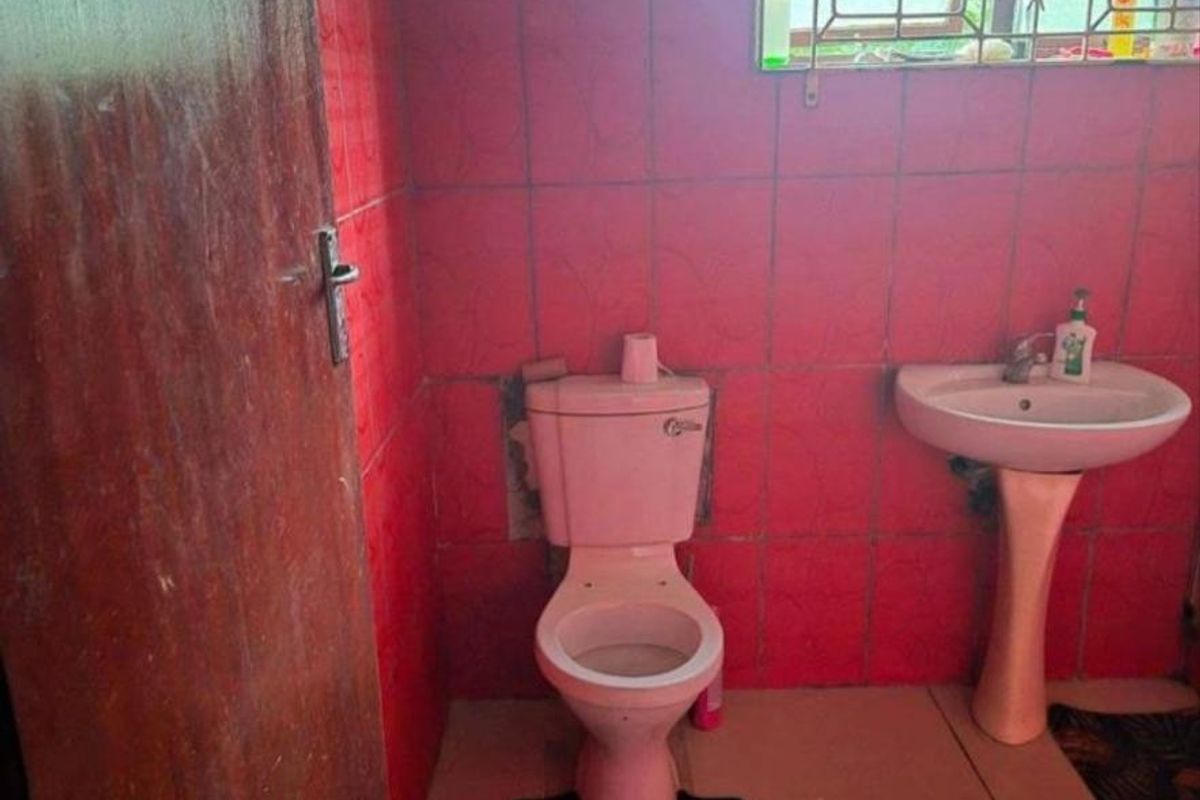 A dated bathroom featuring red wall tiles, a pedestal basin, and a toilet with security bars on the window.