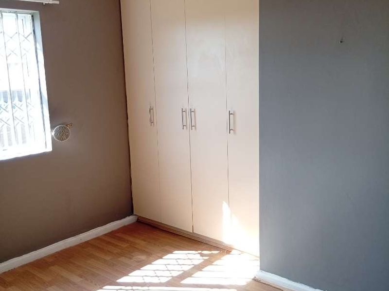 A bright, empty bedroom featuring laminate flooring, built-in cupboards, and a window fitted with security bars.
