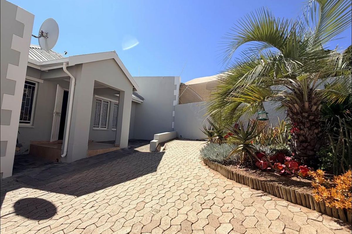 A well-maintained rear exterior area featuring a paved driveway, established garden beds, and clean building facades with security features.