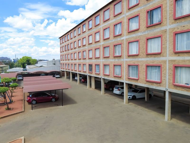 An elevated view of a multi-story brick apartment building featuring covered parking bays and a paved driveway area.