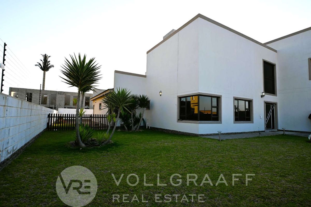 A modern exterior rear view of the property featuring a well-maintained lawn, perimeter wall with electric fencing, and contemporary architectural design.