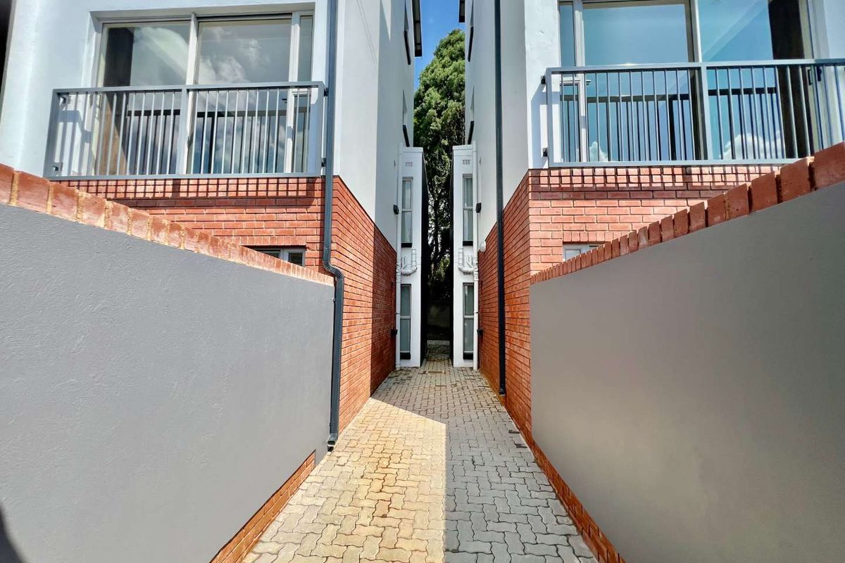 A narrow paved walkway between two modern apartment buildings featuring brick and rendered walls with upper-level balconies.