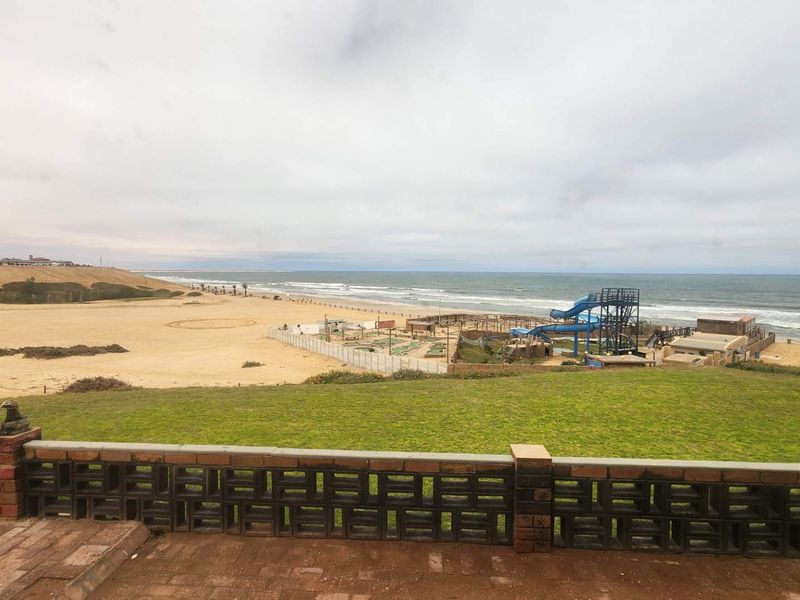 A scenic coastal view overlooking the beach, dunes, and nearby recreational facilities from a paved patio area with a brick boundary wall.