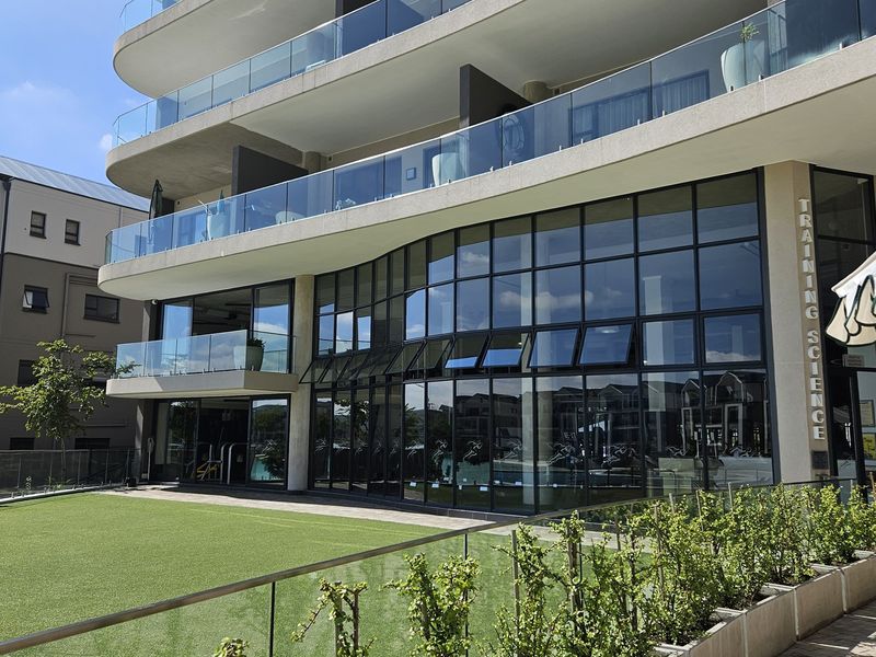 Modern apartment building exterior featuring large glass windows, sleek concrete balconies with glass balustrades, and a well-maintained artificial lawn area.