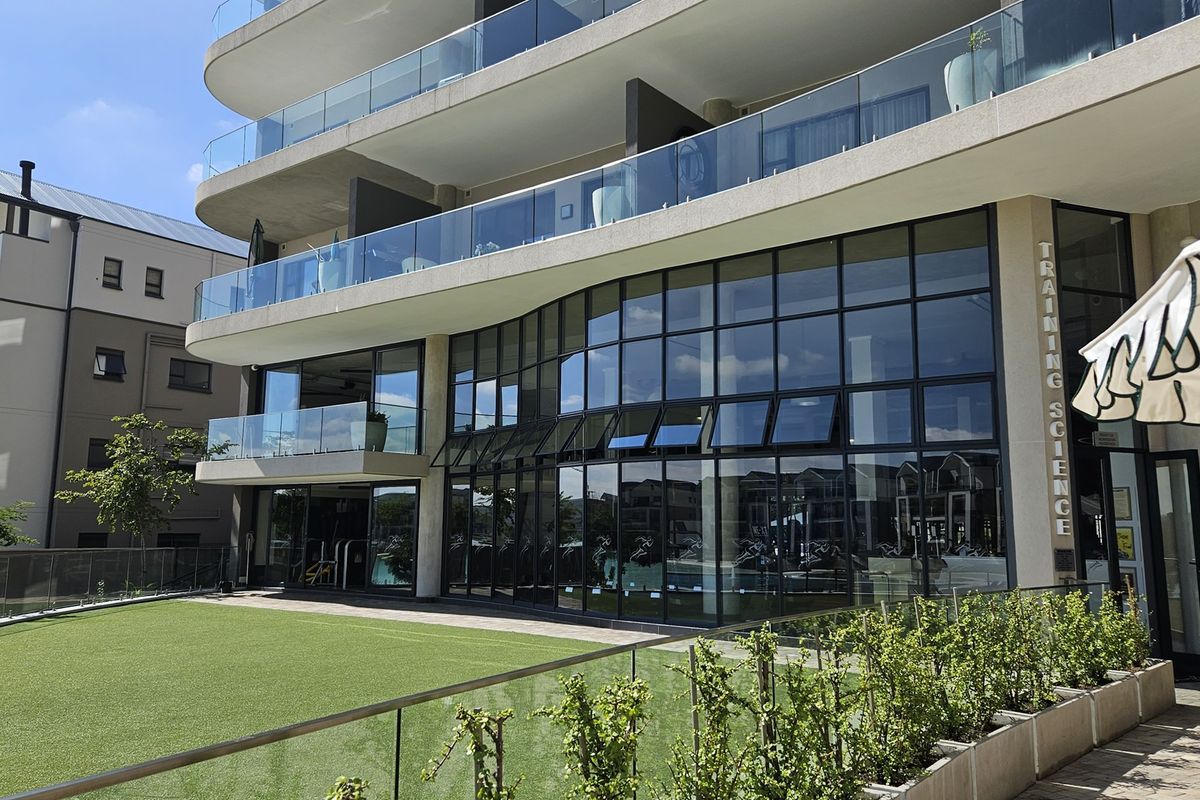 Modern apartment building exterior featuring large glass windows, sleek concrete balconies with glass balustrades, and a well-maintained artificial lawn area.