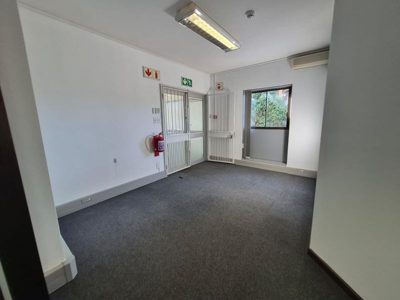 A clean, empty office space featuring carpeted flooring, a split-system air conditioner, security gates, and integrated fluorescent lighting.